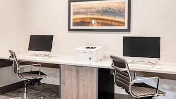 A conference room with two desks, each with a computer monitor, keyboard, and mouse. There is a printer on the central desk. The room has a carpeted floor and a framed picture on the wall.