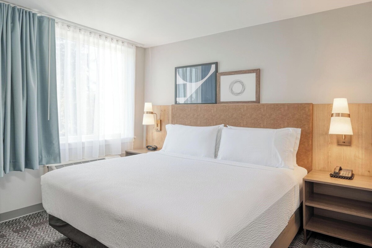 A hotel room with a large bed, two bedside tables with lamps, a headboard, and a window with curtains.