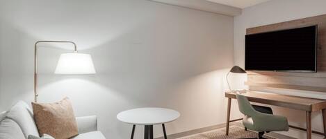 A modern hotel room with a sofa, a small round table, a desk with a chair, and a mounted TV.
