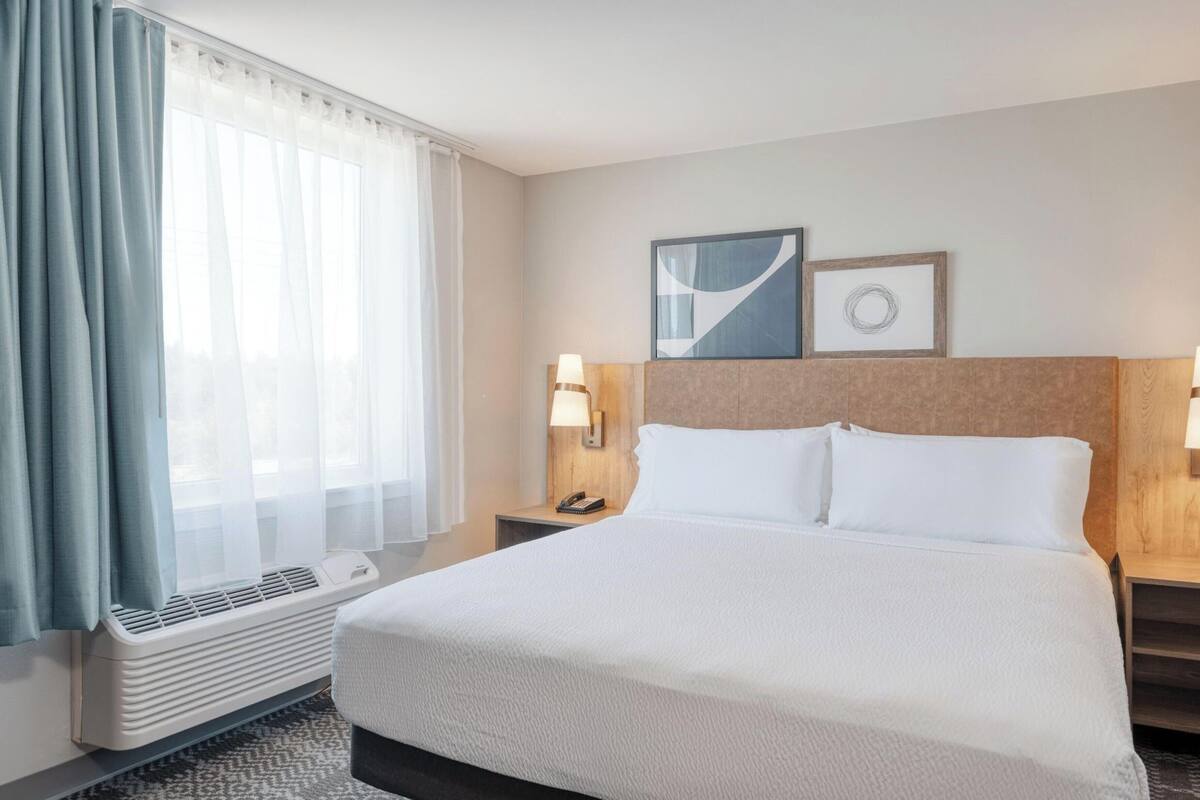 A hotel room with a large bed, a heater, two bedside lamps, a window with sheer curtains, and two framed pictures on the wall.