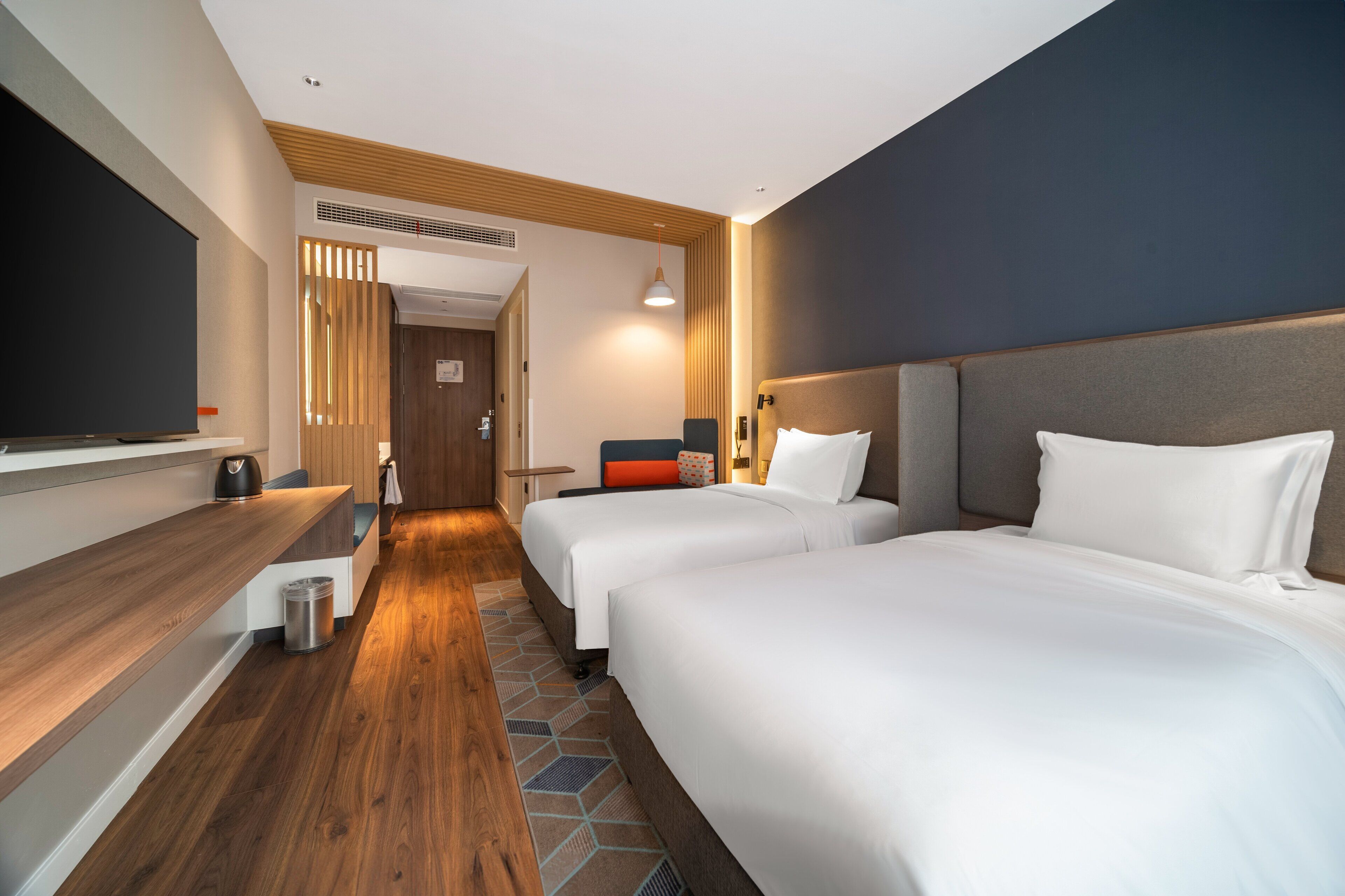 A hotel room with two beds, a wooden floor, a flat-screen TV, and a small kitchenette.