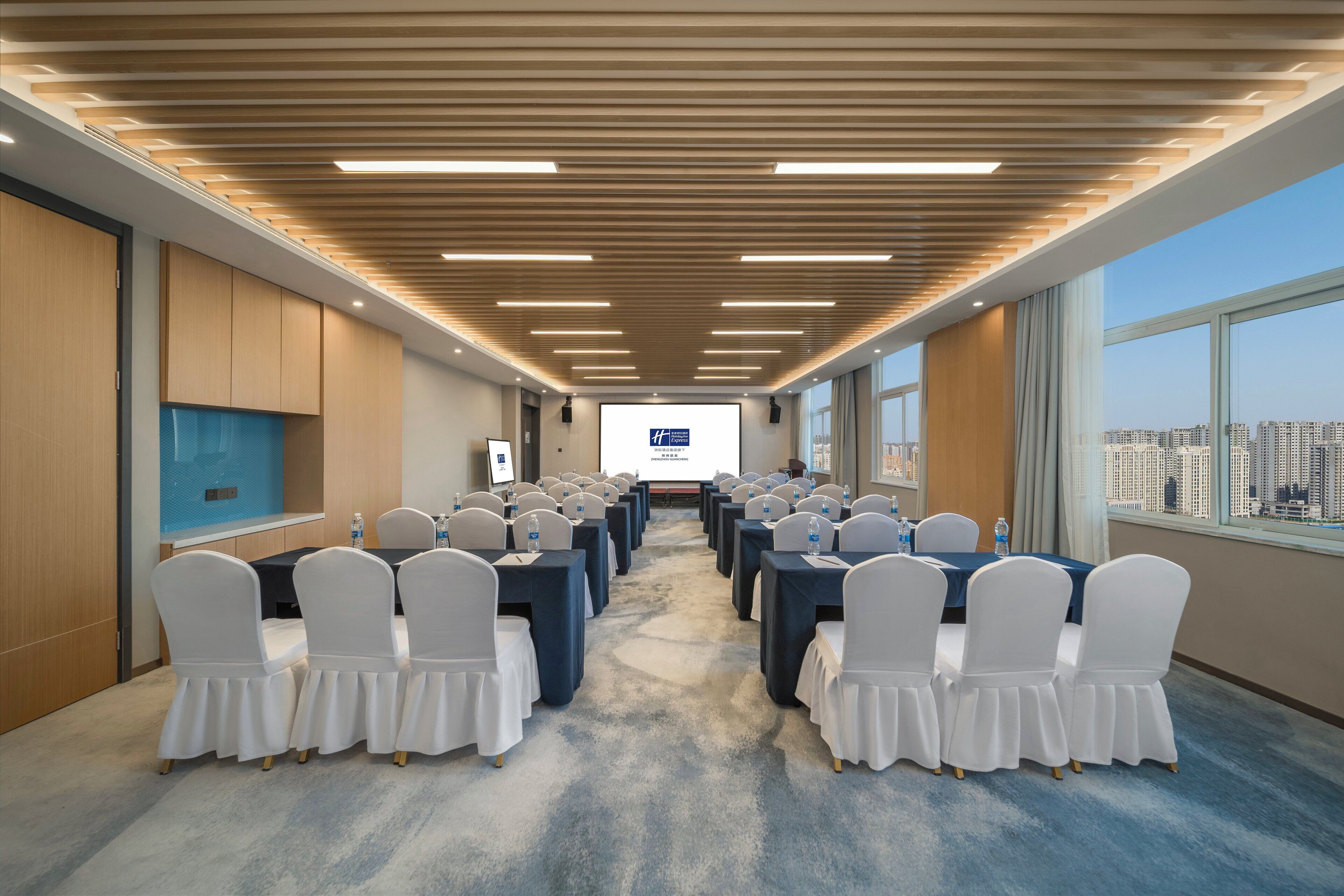 A conference room with rows of tables and chairs, a large screen at the front, and a city view through large windows.