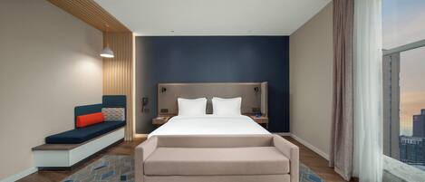 Una habitación de hotel moderna con cama, zona de estar con banqueta y cojines, mesita de noche y vista a la ciudad a través de amplias ventanas.