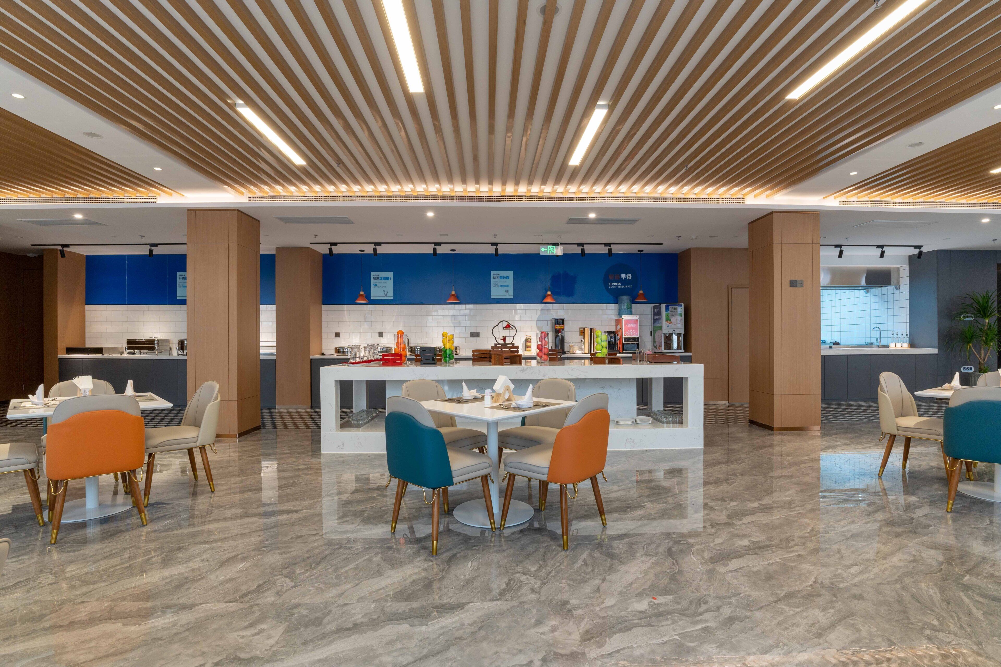 Un moderno vestíbulo de hotel con una mesa redonda central y sillas, una zona de cocina al fondo, e iluminación lineal en el techo.