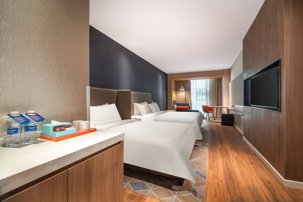 Ein Hotelzimmer mit zwei Betten, einem Fernseher, einem Schreibtisch mit Pflegeprodukten und Blick auf die Stadt.