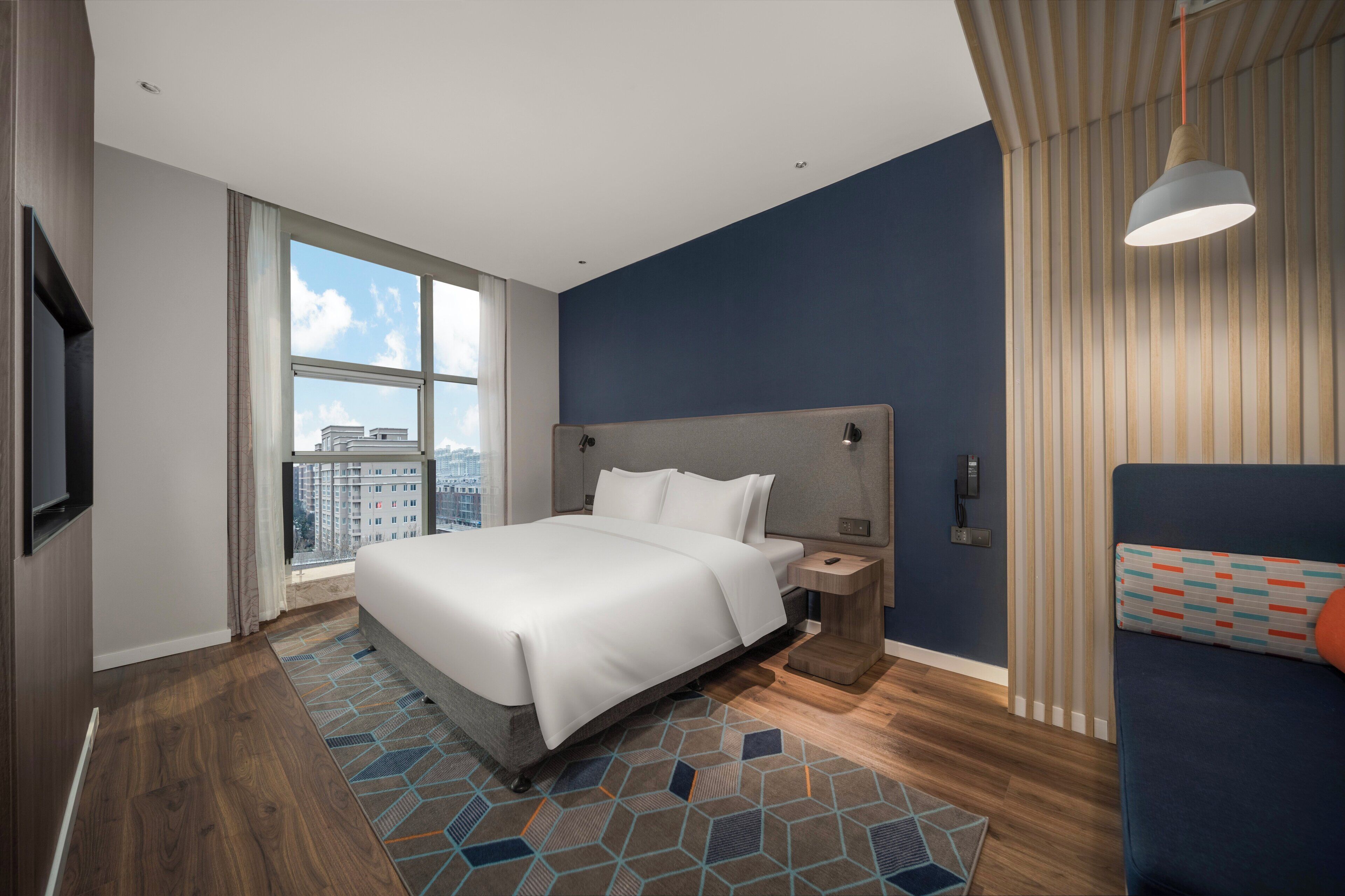 Una habitación de hotel moderna con una cama grande, una mesita de noche, un televisor y vistas a la ciudad.