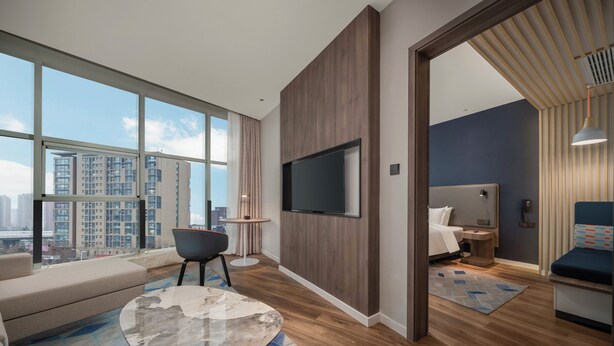 Una habitación de hotel moderna con un ventanal grande, un televisor de pantalla plana, un sofá, una mesa de centro y una cama con cabecera azul.