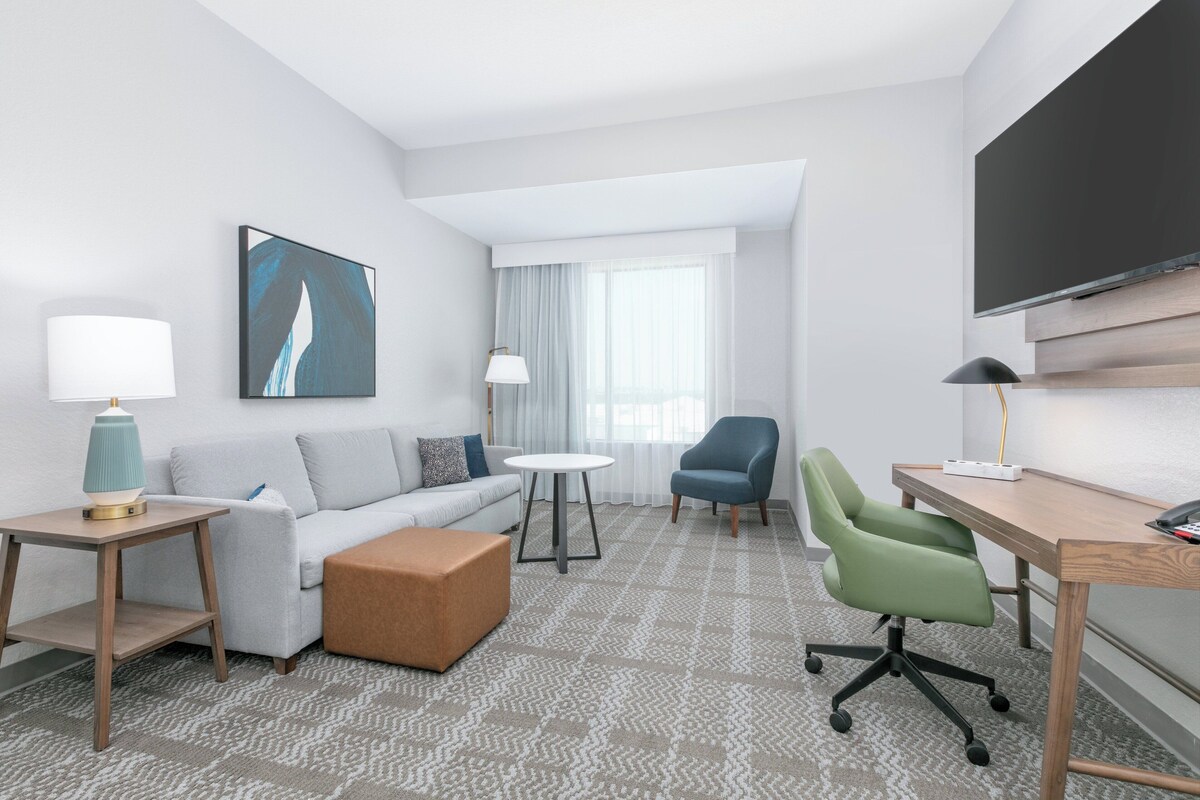 A modern hotel room with a grey sofa, a small round table, a green office chair, a wooden desk, and a large flat-screen TV.