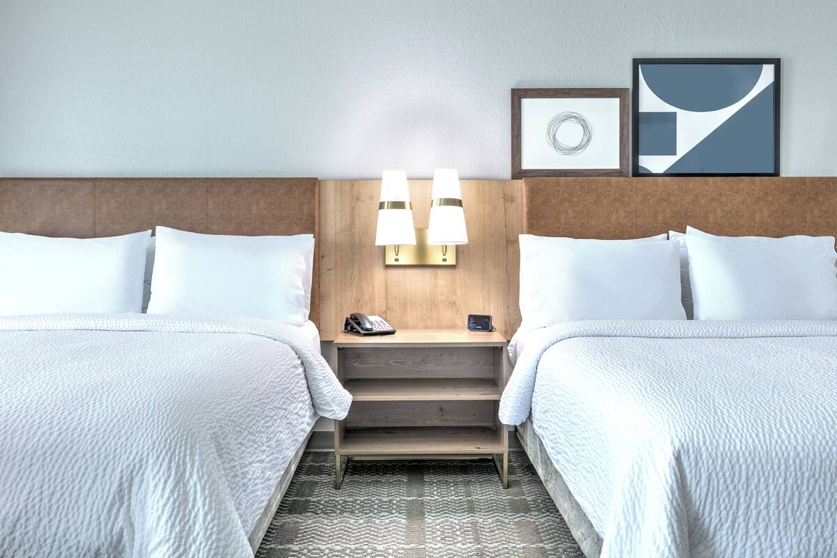 Ein Hotelzimmer mit zwei Betten, einem Holzkopfende, einem Nachttisch mit Telefon und Lampe sowie zwei gerahmten Kunstwerken an der Wand.
