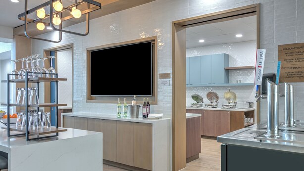 A modern kitchen with a large flat-screen TV, a glass display case with wine glasses, and a breakfast buffet sign. - Staybridge Suites San Antonio Lackland AFB Area by IHG (San Antonio)