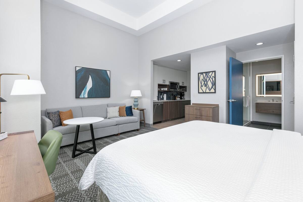 Ein modernes Hotelzimmer mit einem großen Bett, einer Couch, einem kleinen Tisch, einer Küchenzeile und einem durch eine offene Tür einsehbarer Badezimmer.