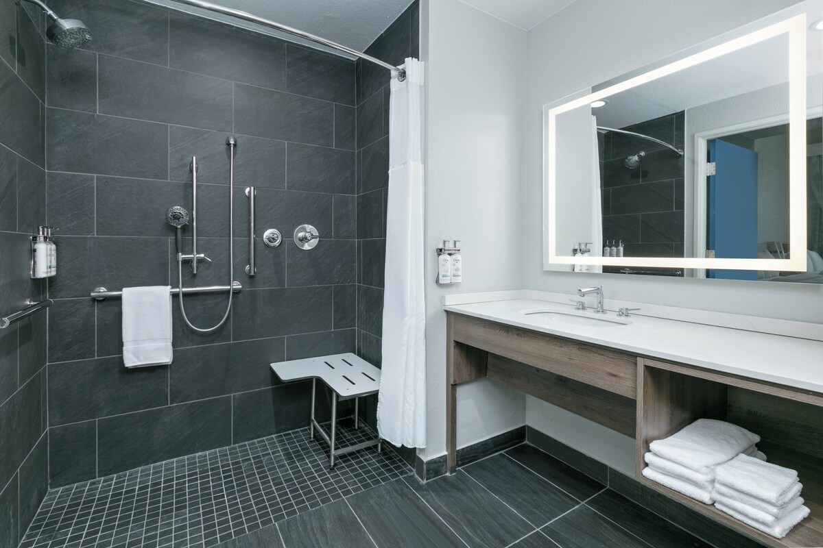 A modern bathroom with a dark tiled shower, a white sink with a wooden vanity, and a large mirror.