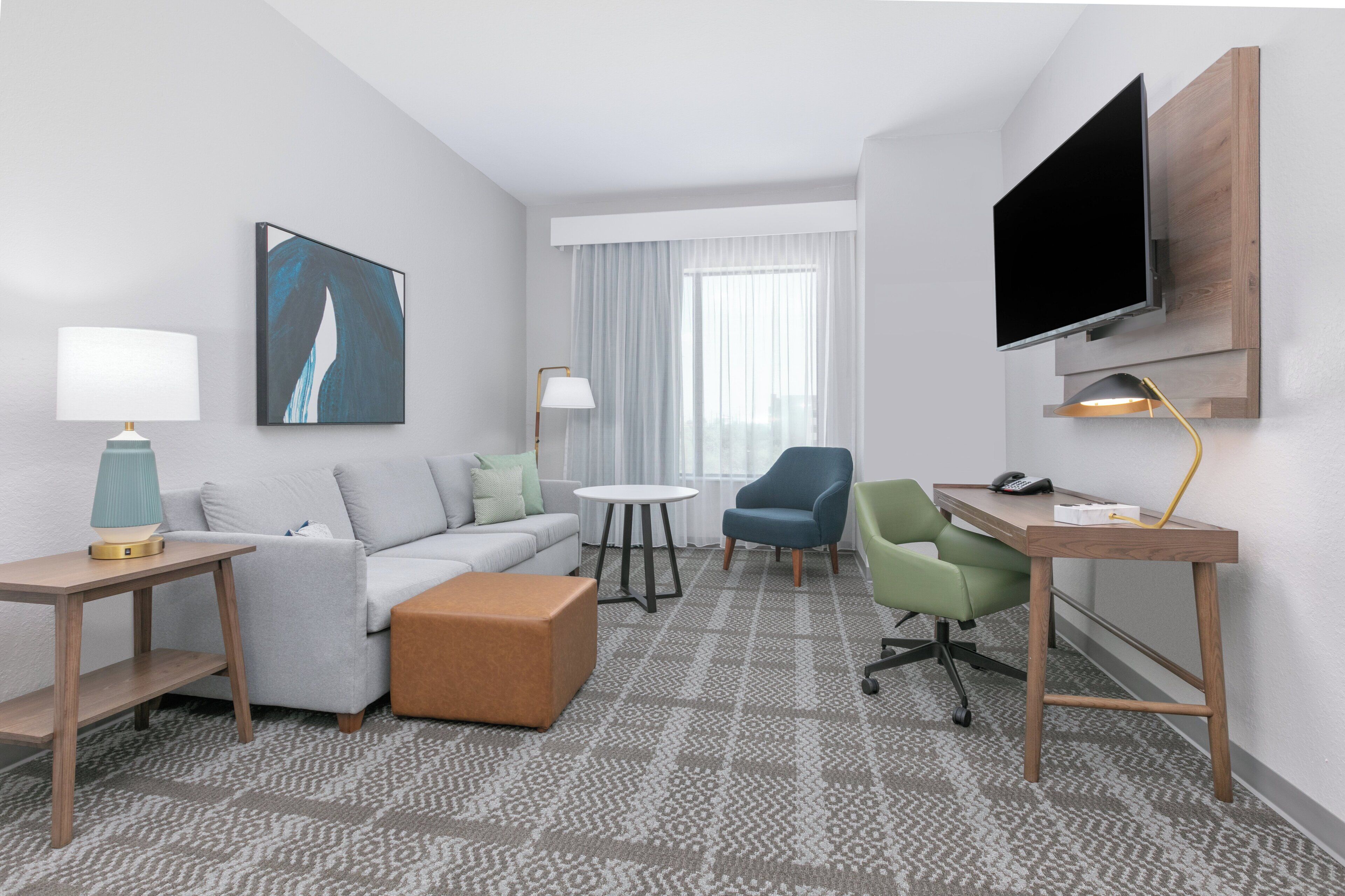 Een moderne hotelkamer met een grijze bank, een klein rond tafeltje, een blauwe fauteuil, een bureau met een computer, een aan de muur bevestigde televisie en een kamerbreed tapijt met een patroon.