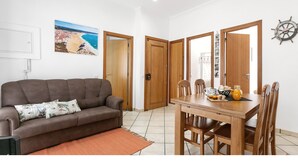Una sala de estar con un sofá marrón, una mesa de comedor de madera con sillas, una alfombra y un cuadro de una playa.