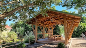 Un pabellón de madera con bancos y una mesa, rodeado por un sendero de grava y follaje.
