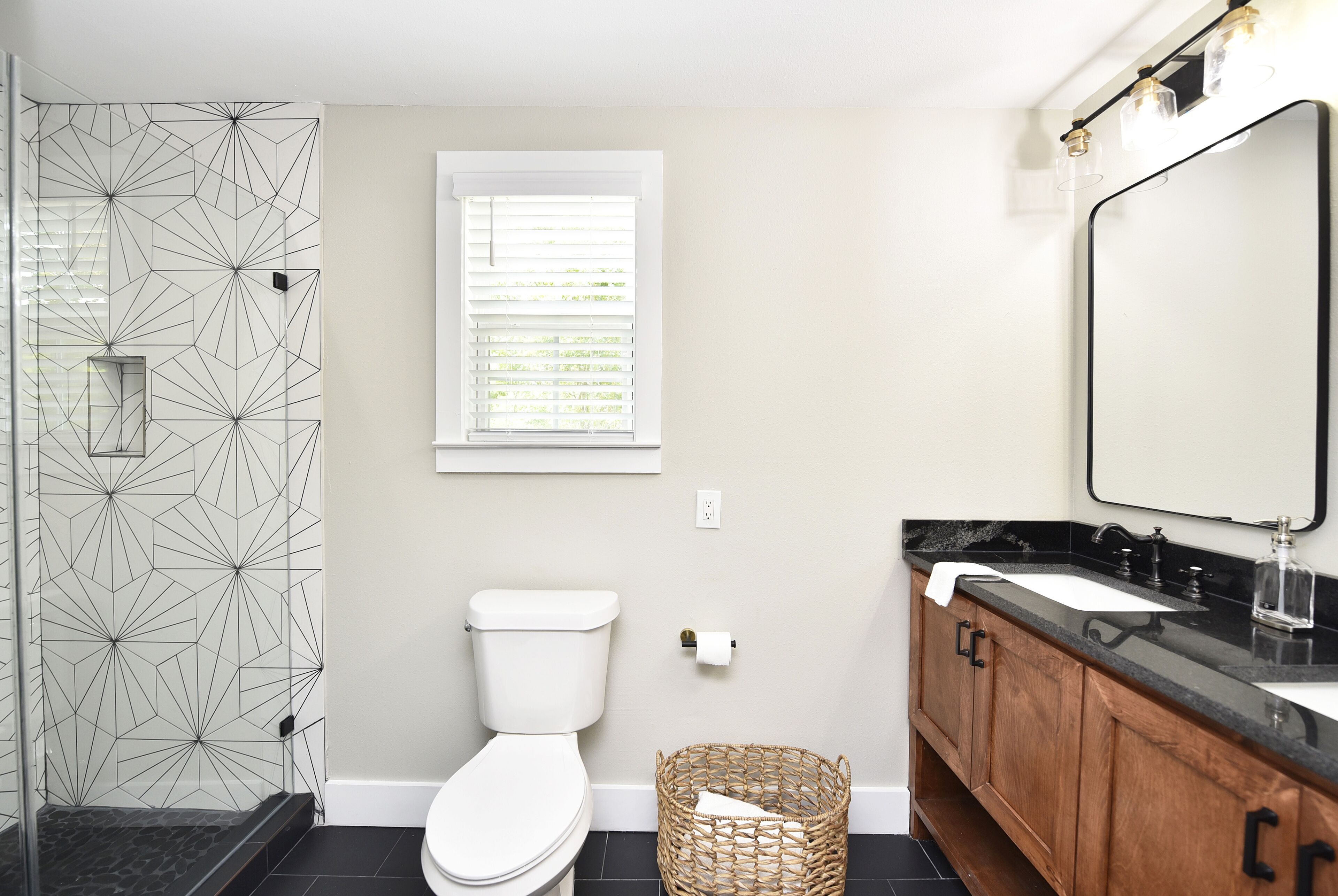 A bathroom with a geometric-patterned shower wall, a toilet, a window with blinds, a wooden vanity with a black countertop, and a mirror with lights above it.