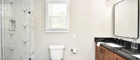 A bathroom with a geometric-patterned shower wall, a toilet, a window with blinds, a wooden vanity with a black countertop, and a mirror with lights above it.