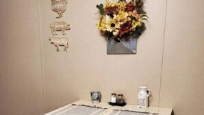Et lille spisebord med en servietring, to stole, et dekorativt vægophæng og en lille vase med tørrede blomster.