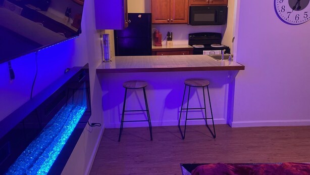 A compact kitchen with wooden cabinets, a black refrigerator, a microwave, and a clock on the wall. - Cozy apartment steps from Christ Hospital, UC and minutes to downtown! (Cincinnati)