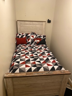 A small, narrow room with a wooden bed frame and a geometric-patterned bedspread.