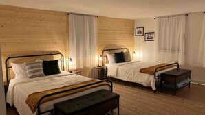 Une chambre avec deux lits, chacun doté d’une tête de lit en bois, d’une literie blanche et d’un banc au pied de l’un des lits. On y trouve des tables de chevet avec des lampes, un tapis sur le sol et un tableau accroché au mur.