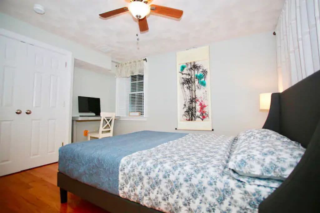 A bedroom with a bed, a desk, a chair, a ceiling fan, and a wall-mounted TV.