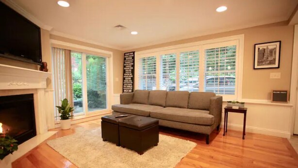 A living room with a fireplace, a sofa, a coffee table, and a large window with blinds. - Peaceful 2BR near US Route 1 and Boston. (Melrose)