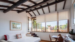 A room with a bed, a sofa, a dining table, and chairs. There is a window with a view of greenery outside. - Valeria Terrace   Monti, Rome (ROMA)