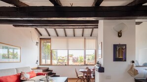 A living room with a sofa, a coffee table, and a dining area with chairs and a table. There is a window with a view of the outdoors. - Valeria Terrace   Monti, Rome (ROMA)