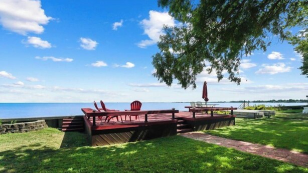 A lakeside deck with red chairs and a table, surrounded by grass and a stone wall. - Restored historic log cabin and deck on Lake Erie (Luna Pier)