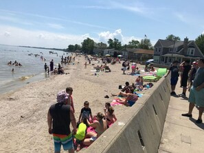 A beach with people sunbathing and swimming, a concrete barrier, and houses in the background. - Restored historic log cabin and deck on Lake Erie (Luna Pier)
