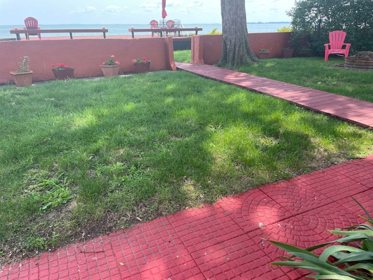 A grassy area with a red brick pathway leading to a seating area with chairs and a table.