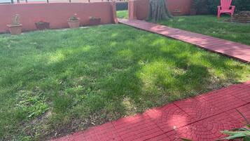 A grassy area with a red brick pathway leading to a seating area with chairs and a table.