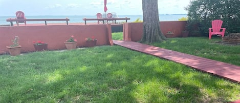 Un espacio con césped y un sendero de ladrillo rojo que conduce a un área de descanso con sillas y una mesa.
