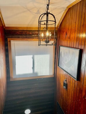 A wooden-paneled hallway with a chandelier, a window with blinds, and a framed picture on the wall. - Restored historic log cabin and deck on Lake Erie (Luna Pier)