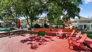 A wooden deck with red chairs and a red umbrella, surrounded by trees and houses. - Restored historic log cabin and deck on Lake Erie (Luna Pier)