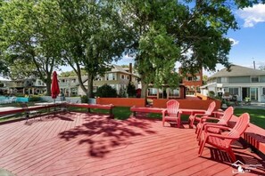 A wooden deck with red chairs and a red umbrella, surrounded by trees and houses. - Restored historic log cabin and deck on Lake Erie (Luna Pier)