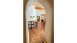 Visão através de uma porta arqueada para uma área de refeições com piso ladrilhado e teto de madeira.