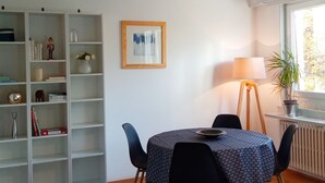 Un comedor con una mesa, sillas, una estantería, un cuadro enmarco, una lámpara y una ventana con una planta.