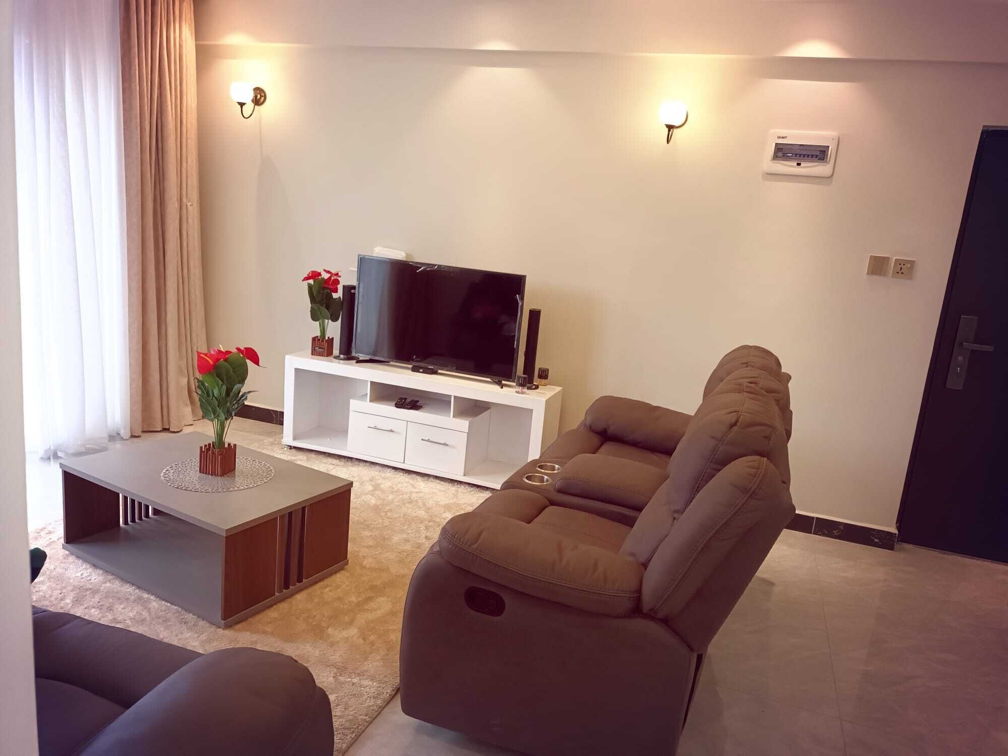 Een moderne woonkamer met een verstelbare bank, een flatscreen televisie op een witte tv-meubel en een kleine salontafel met een vaas rode bloemen.