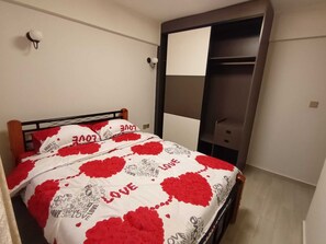 Une chambre à coucher avec un lit recouvert d’une couvre-lit blanche et rouge à motifs de cœurs, une tête de lit en bois et une armoire avec un miroir.