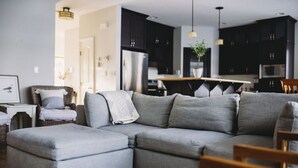 A modern living room with a grey sectional sofa, a small ottoman, a wicker chair, and a dining area with a wooden table and chairs. - The Lakehouse (Lloydminster)