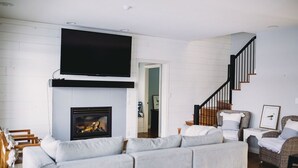 A living room with a fireplace, a large flat-screen TV, a staircase, and wicker furniture. - The Lakehouse (Lloydminster)