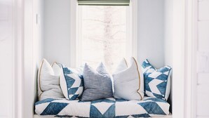 A cozy seating area with a bench, patterned cushions, a window with blinds, and a hanging light fixture. - The Lakehouse (Lloydminster)