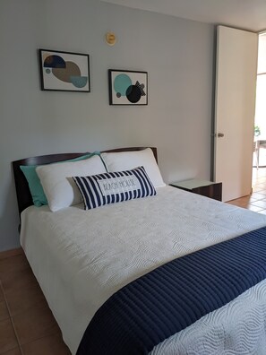 A neatly made bed with a white and navy blue quilt, a pillow labeled "BEACH HOUSE", and two abstract wall art pieces.