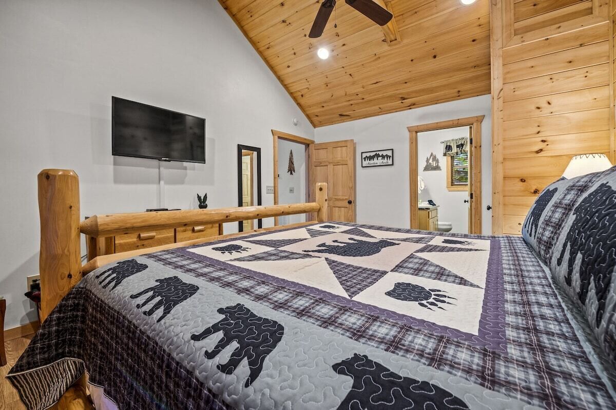 A bedroom with a large bed, wooden ceiling, a mounted TV, and a wooden railing.