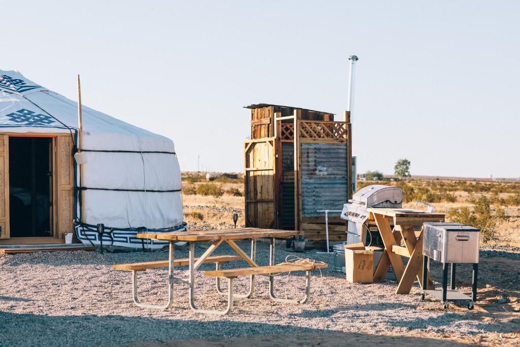 Une yourte équipée d’une table de pique-nique et d’un barbecue, entourée d’une structure en bois et d’des toilettes portables.