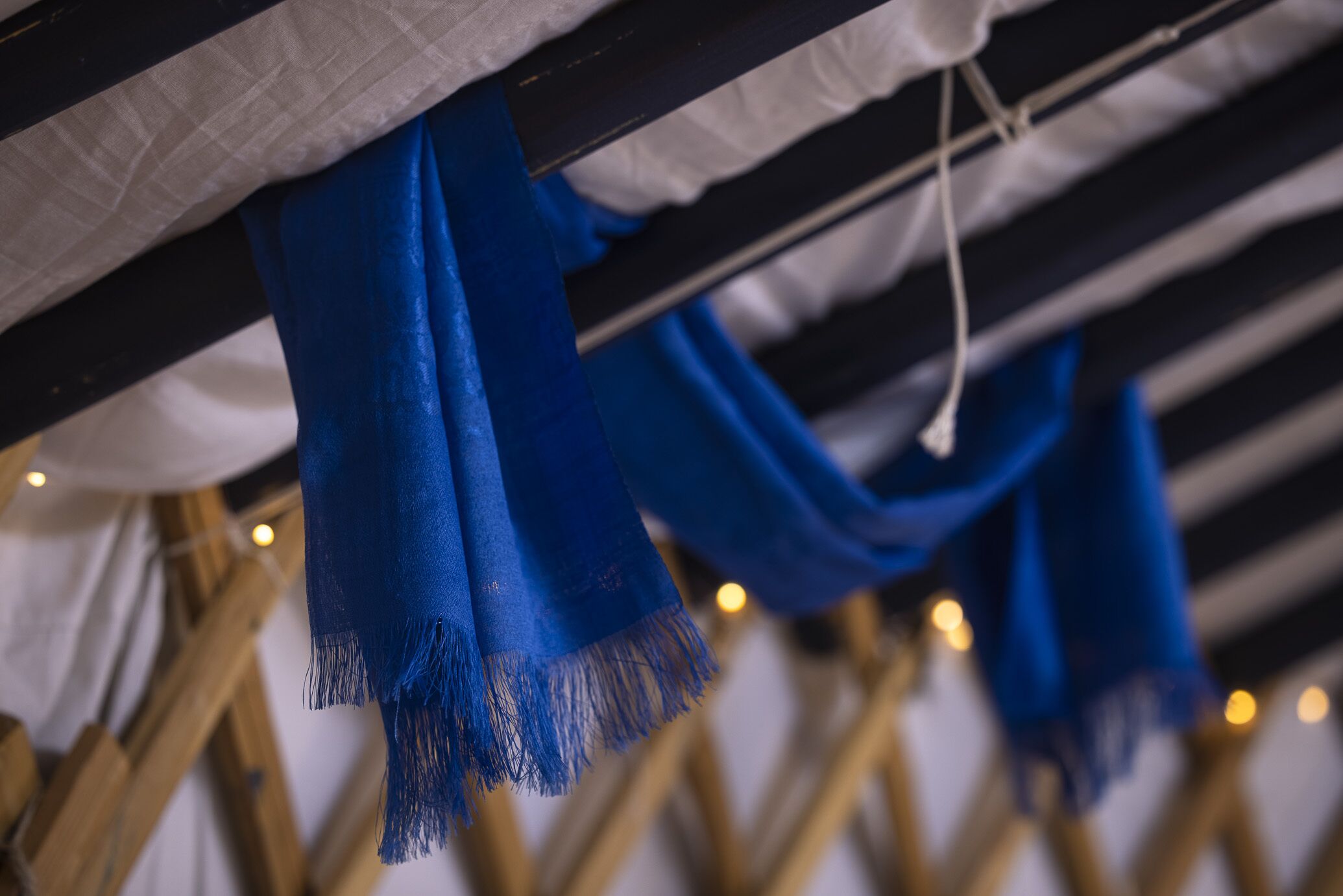 A room with a bed covered in white linens, blue scarves hanging from the headboard, and a wooden frame structure.