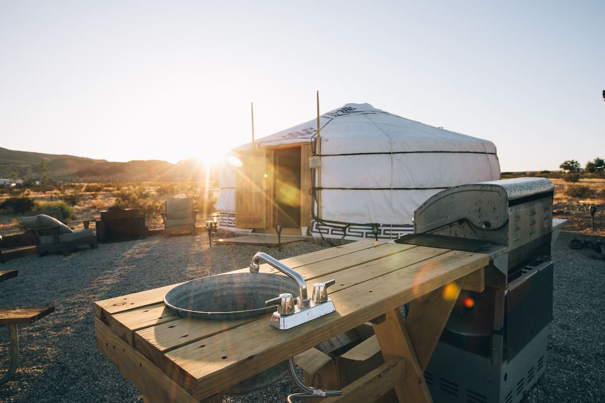 A wooden picnic table with a sink and a grill, set against a backdrop of a yurt and a clear sky.