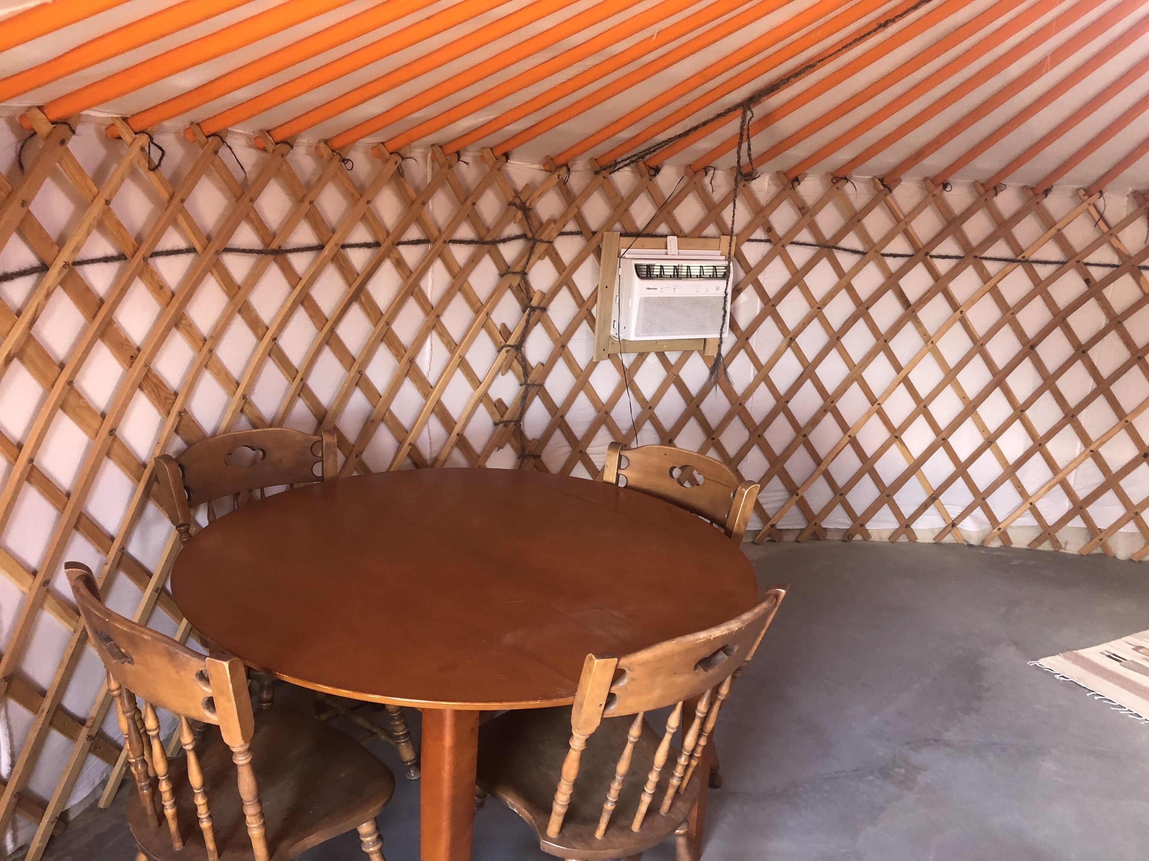 A round wooden table with four chairs, a white air conditioning unit, and a wooden lattice wall.